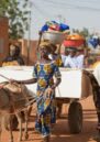 A procession of wedding guests walk along with a donkey and cart on an earthen red road with gifts and baskets.