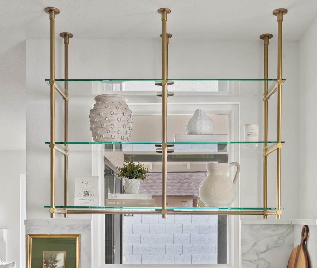 Ceiling-mounted glass shelving in brushed brass suspended over a marble kitchen counter
