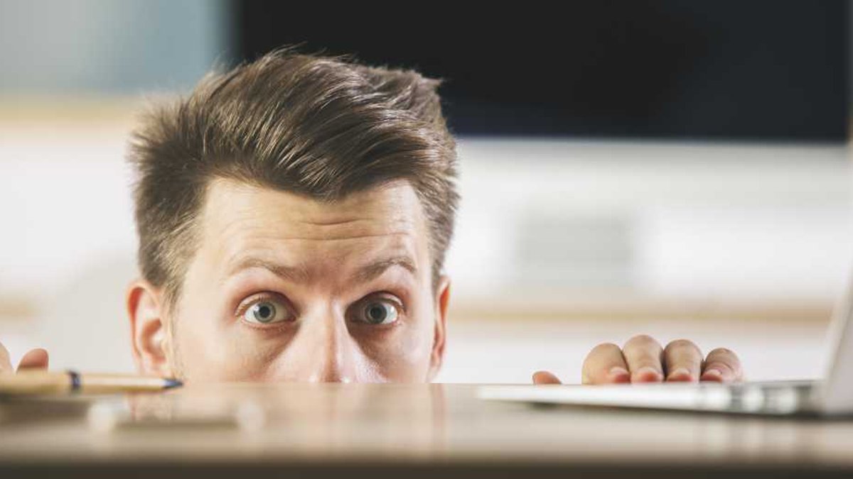 Man hiding behind blurry wooden desktop with laptop. Fearful anxious worker.
