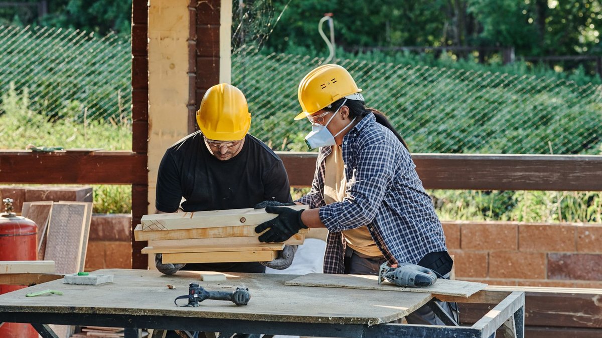 How to Protect Your Renovation Budget if the Builder Fails Two construction workers organize wooden planks on a table at a sunny outdoor site.
