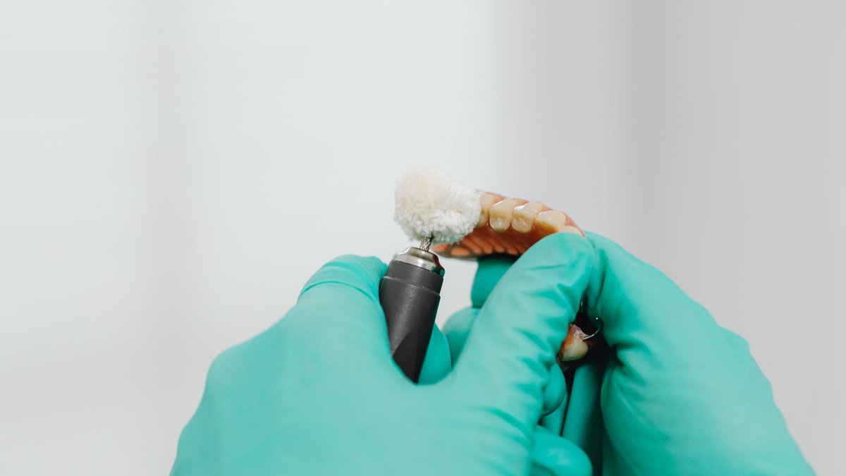 A close-up view of hands with latex gloves polishing a denture using a dental instrument.
