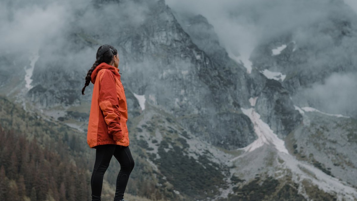 REI discounts hiking boots and shoes Woman in orange jacket hiking through foggy mountain landscape, admiring the view.