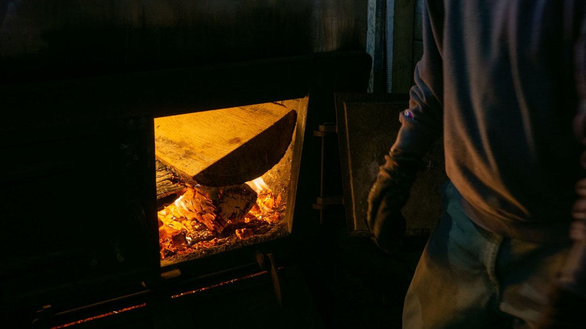 Experts weigh pros and cons of stove fuels Person tending to a wood fire in a dimly lit room, adding logs to the flames.
