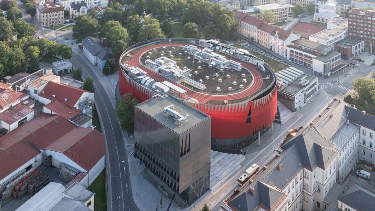 Czechia's Jihlava Arena Revitalizes Urban Life and Culture Aerial view of a modern, oval-shaped red building with a flat rooftop surrounded by traditional city buildings, trees, and ro