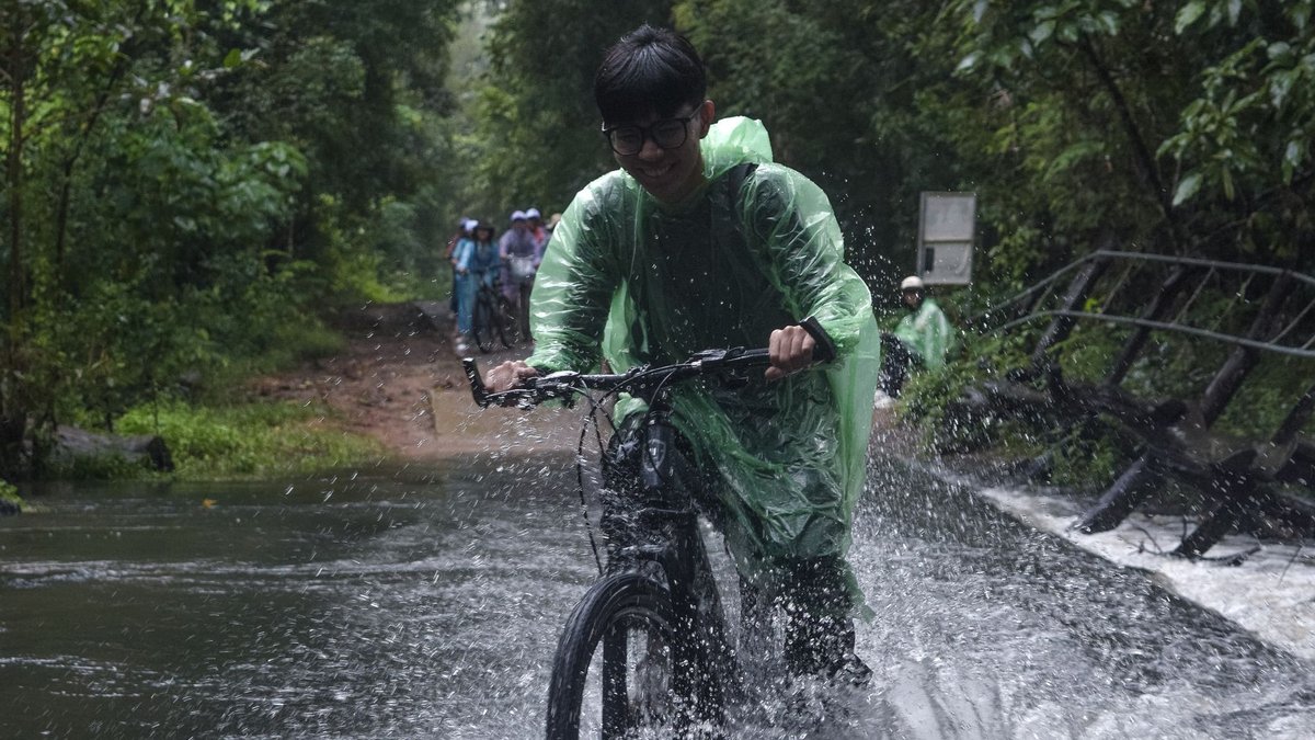 A cyclist crosses a flooded path in a lush rainforest, creating a splash of water under a cloudy sky.