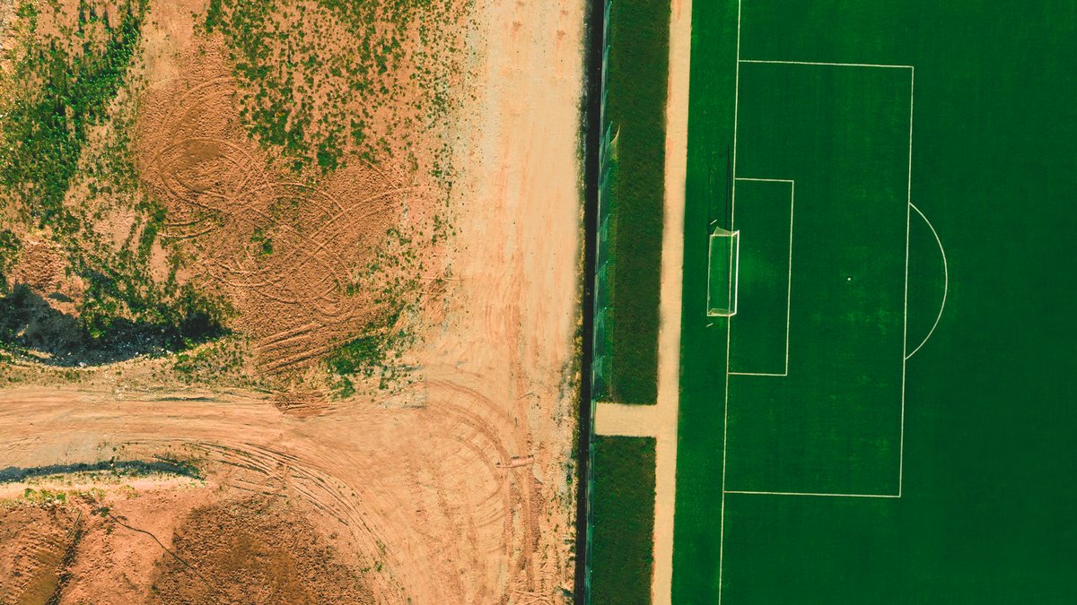 Top-down aerial view of a green football pitch next to a barren dirt field.
