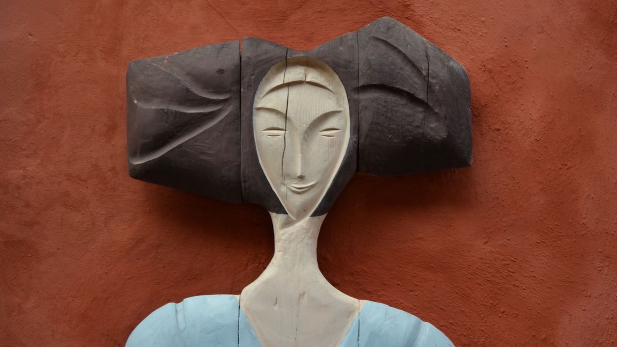Wooden figure in blue attire with abstract hair on terracotta backdrop.