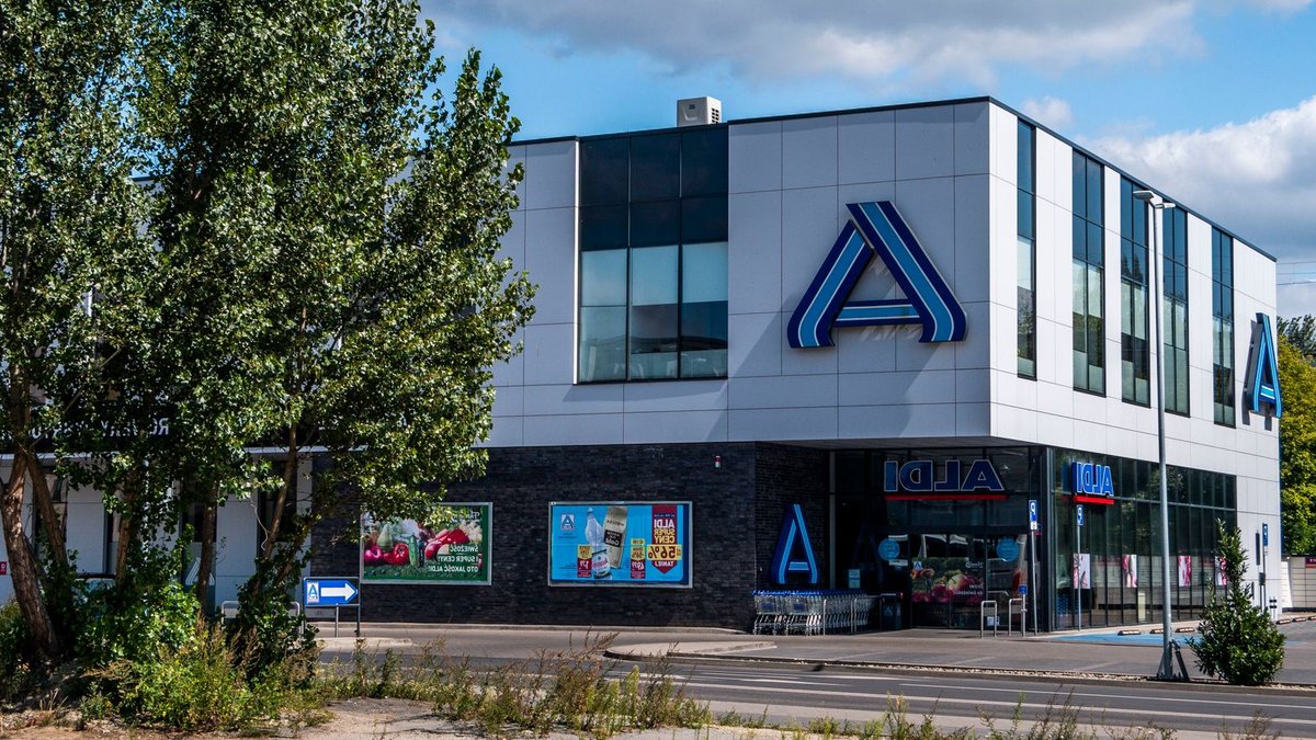Blue and white modern Aldi store exterior surrounded by green trees under a clear sky.