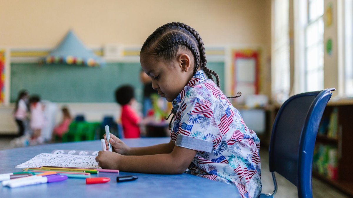 A young child with braids engaged in coloring at a classroom desk surrounded by school supplies.