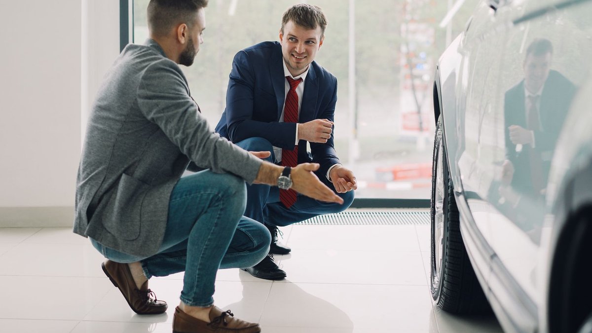 Two men discussing car features in a showroom, kneeling near a vehicle.