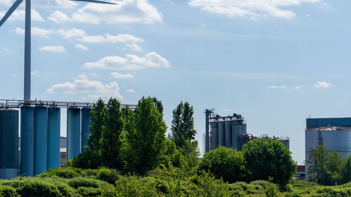 Scenic industrial landscape featuring a wind turbine, factories, and lush greenery by a body of water.