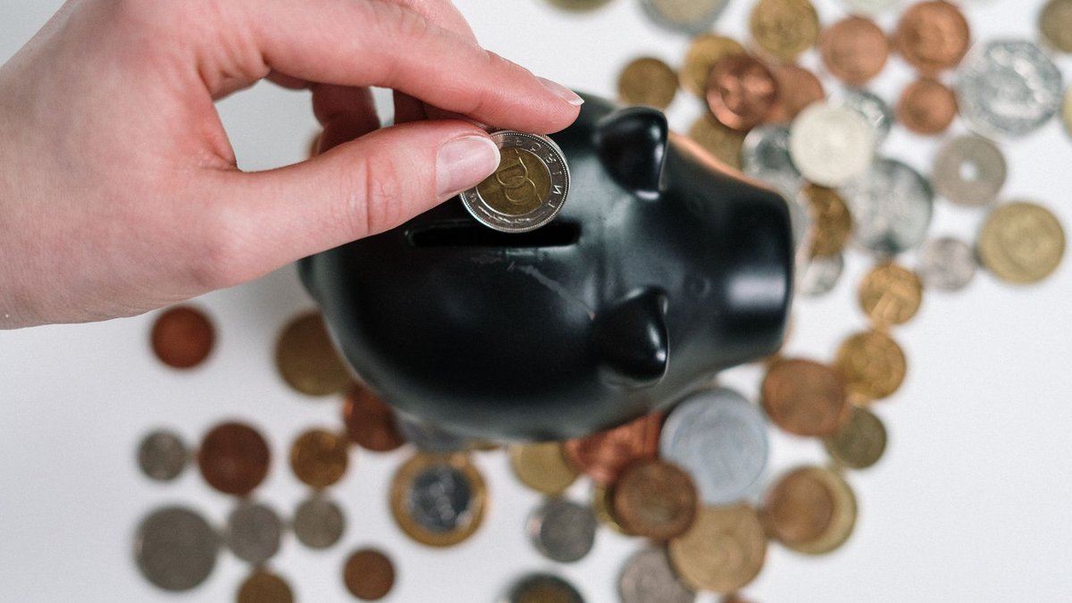Close-up of a hand inserting a coin into a black piggy bank with scattered coins on a white background.