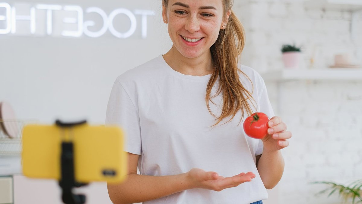 Stricter food safety rules for online sellers A woman demonstrating cooking online with a smartphone, holding a tomato.
