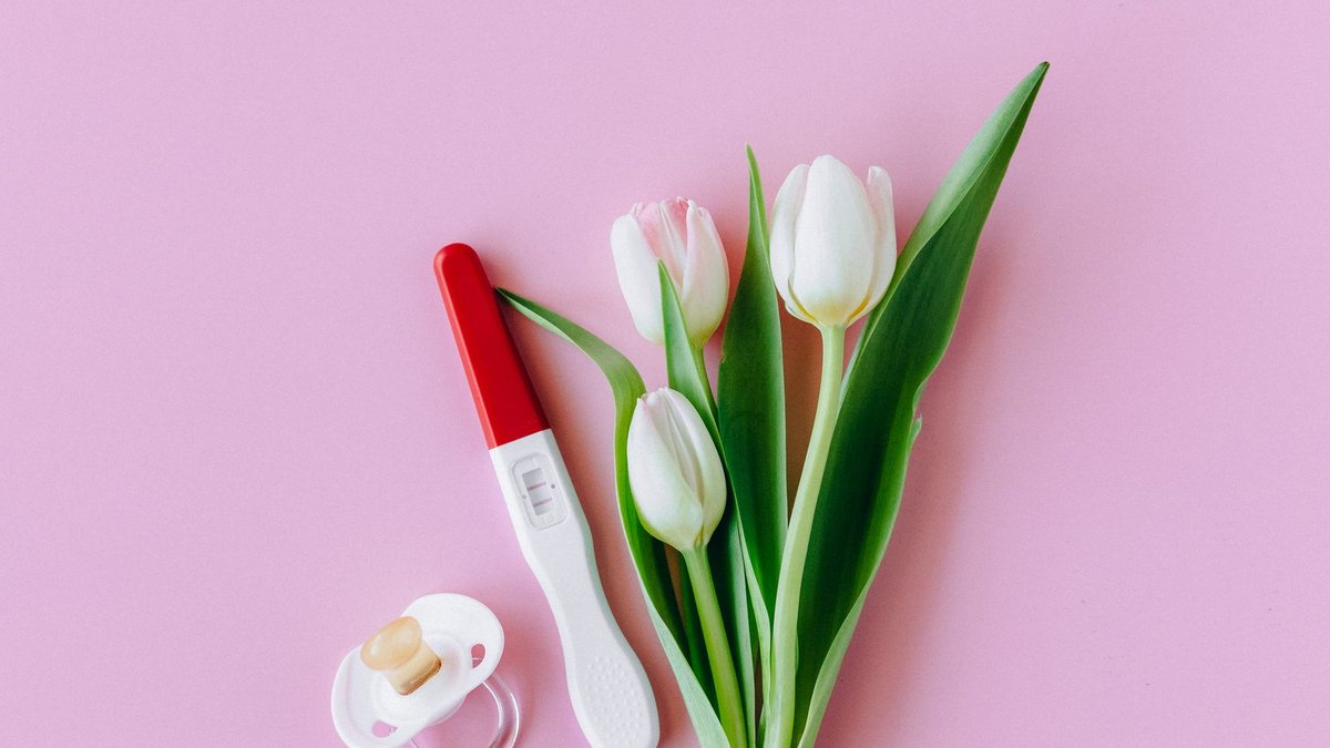Flat lay of pregnancy test, pacifier, and tulips on pink surface conveying fertility and new beginnings.
