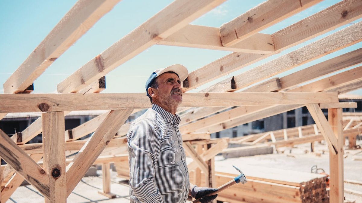 A construction worker surveys a wooden roof frame build on a sunny day.