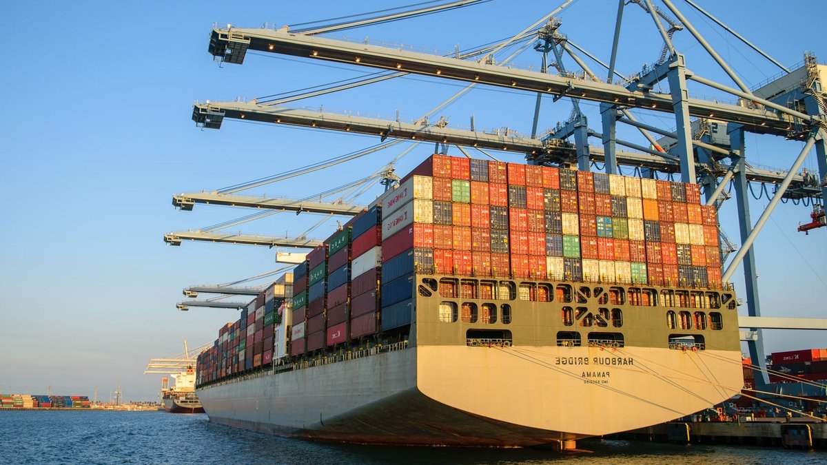 Firefighting Continues on Damaged Espirito Santos Express A large container ship at an industrial harbor with cranes under a clear blue sky.