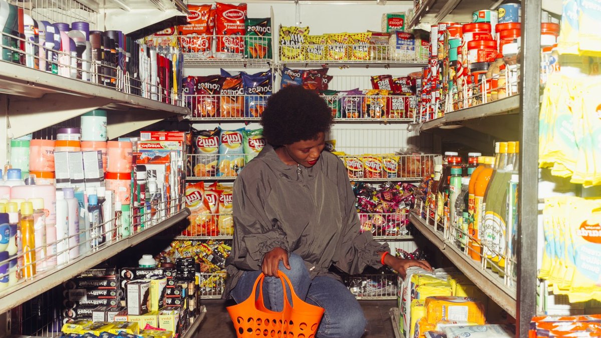 Full length of adult ethnic female buyer selecting goods while squatting with shopping bag among grocery shelves.
