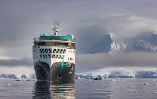 A white and teal expedition cruise ship navigating through icy Antarctic waters, with snow-covered