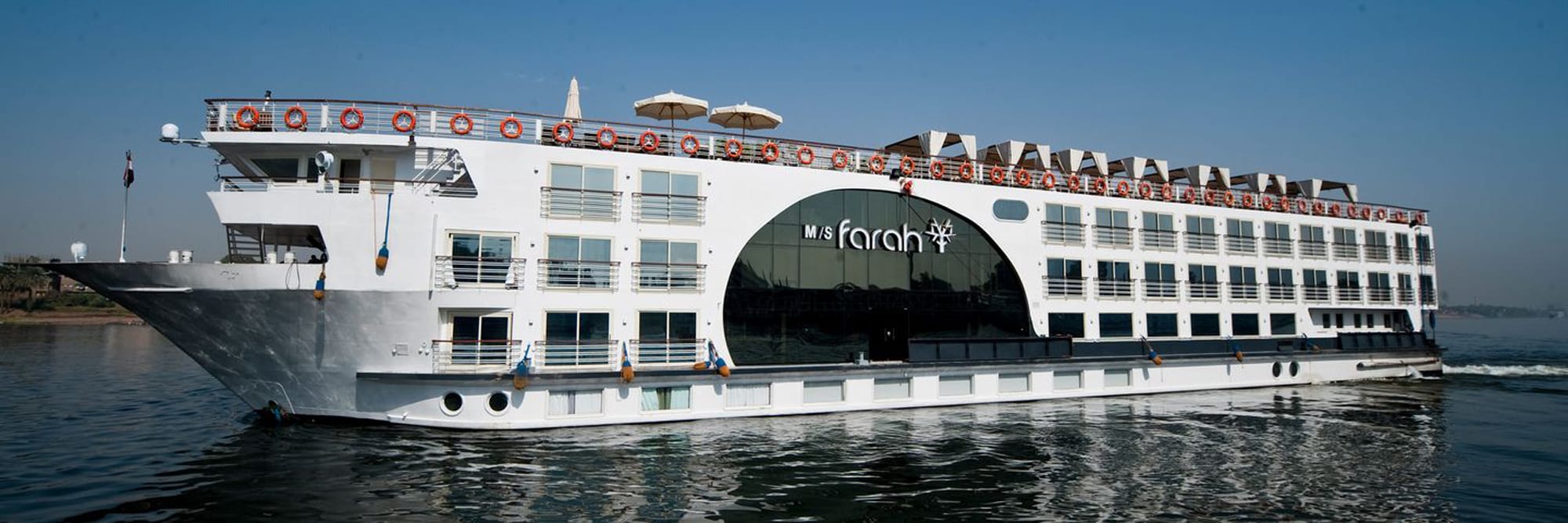 The M/S Farah, a modern multi-deck river cruise ship, is moored on the Nile River. The white vessel