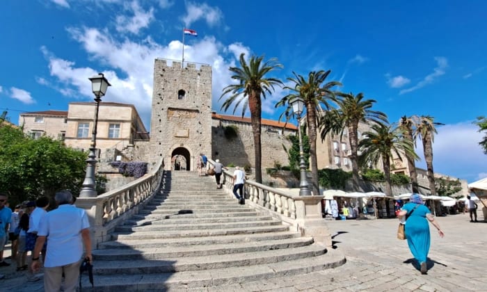 A scenic plaza featuring a medieval stone tower with a flag at its peak, connected by a grand