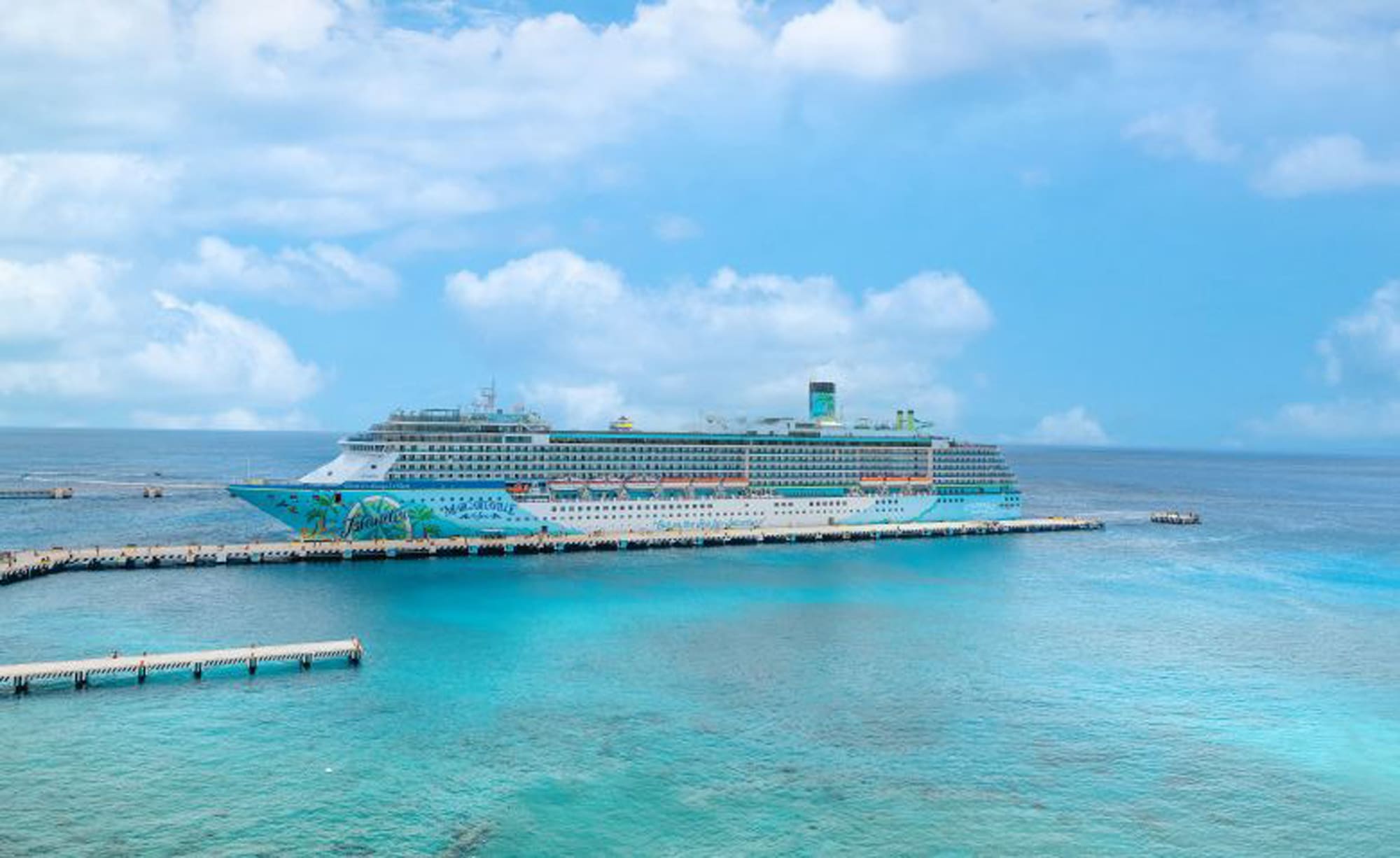 A large, colorfully decorated cruise ship moored at a pier with crystal clear blue-green waters surrounding it. The ship is set against a bright blue sky with fluffy white clouds.