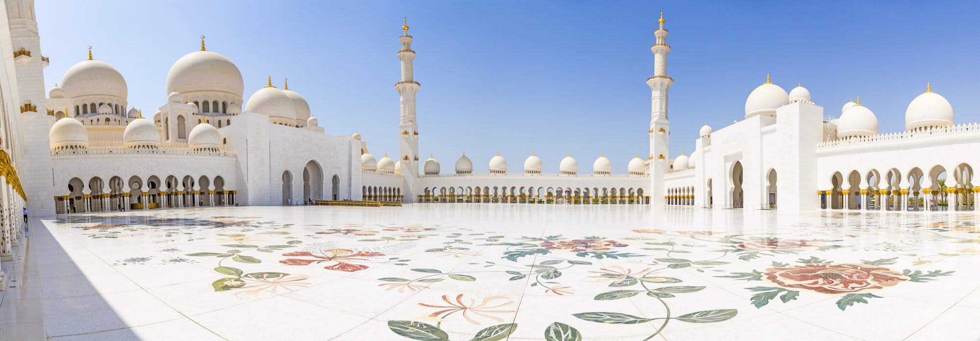 A stunning view of the Sheikh Zayed Grand Mosque in Abu Dhabi, featuring its iconic white marble architecture with golden domes and minarets, ornate floral marble inlay patterns covering the vast courtyard floor, and pristine Islamic geometric design elements under clear blue skies.