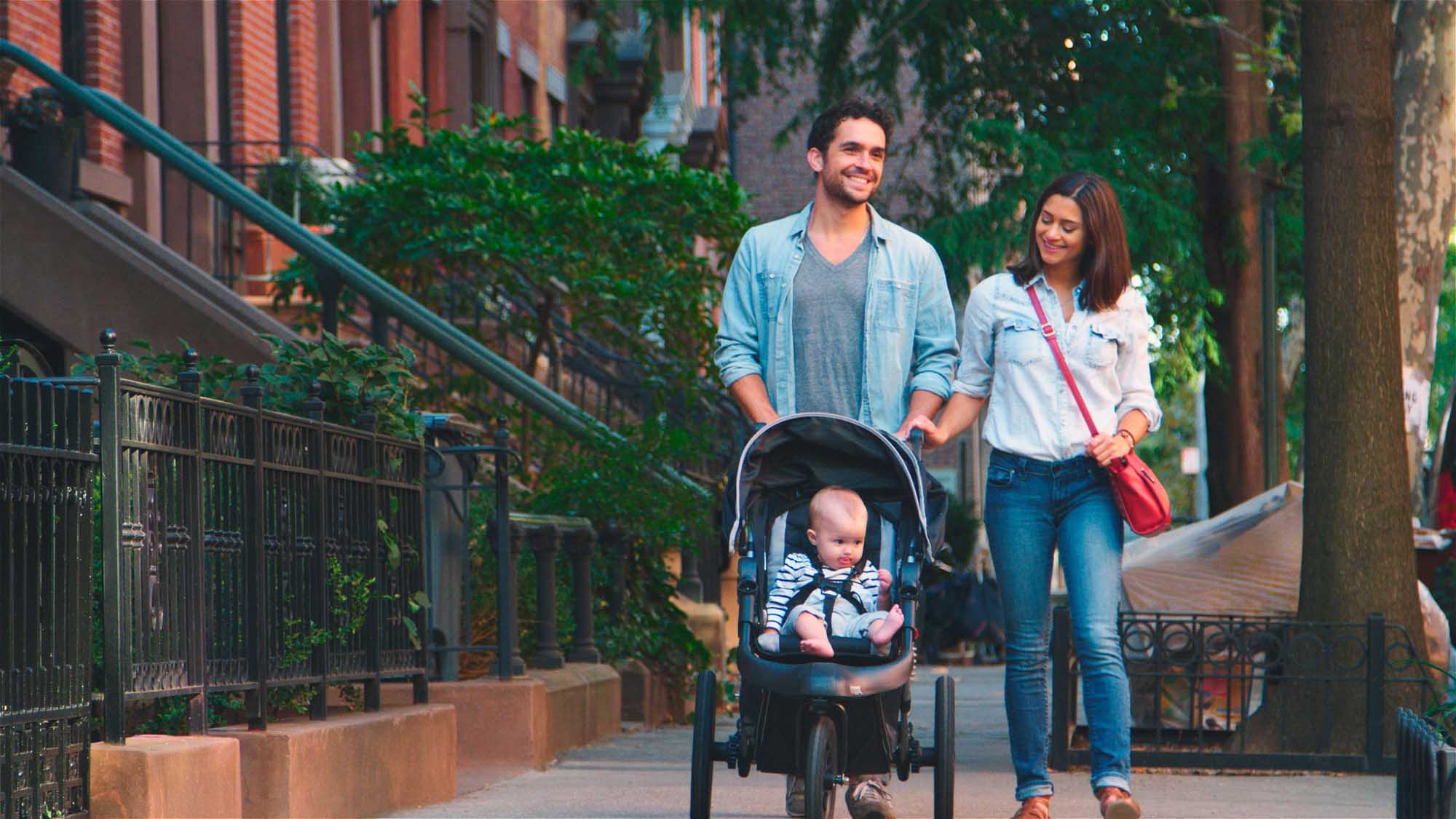 A young family enjoys a leisurely walk through a tree-lined urban street, with the father pushing