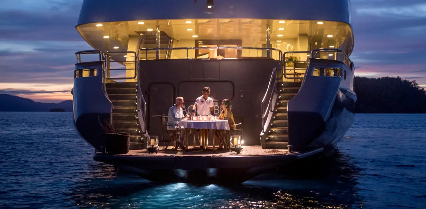 An elegant outdoor dining experience on the illuminated stern platform of a luxury yacht during golden hour, with a family of four seated at a formally set table while the sun sets over calm coastal waters.