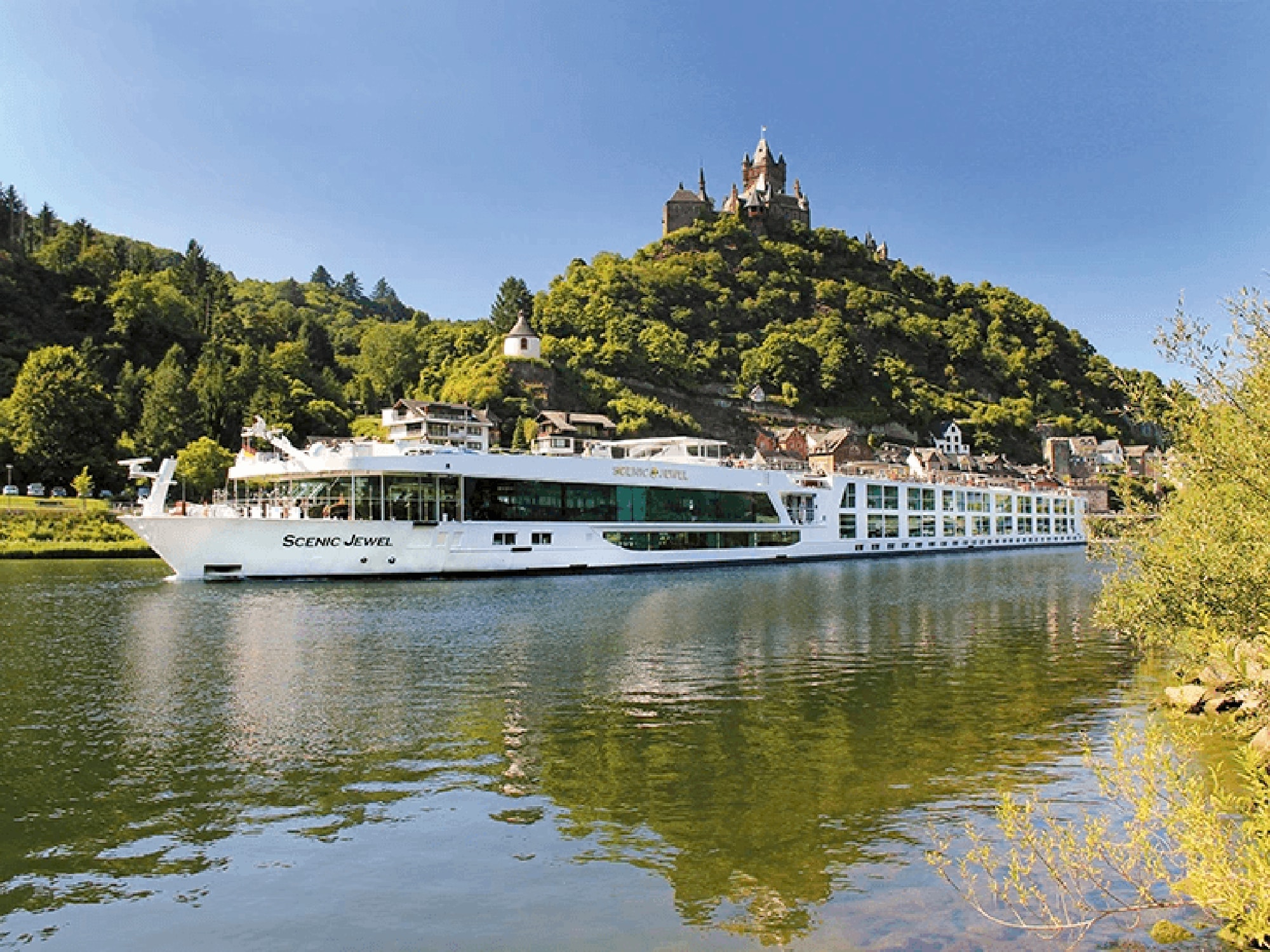 A white river cruise ship named Scenic Jewel sailing on a tranquil river with a historic hilltop castle and forested landscape in the background. The scenic setting showcases the picturesque beauty of a European river cruise destination.