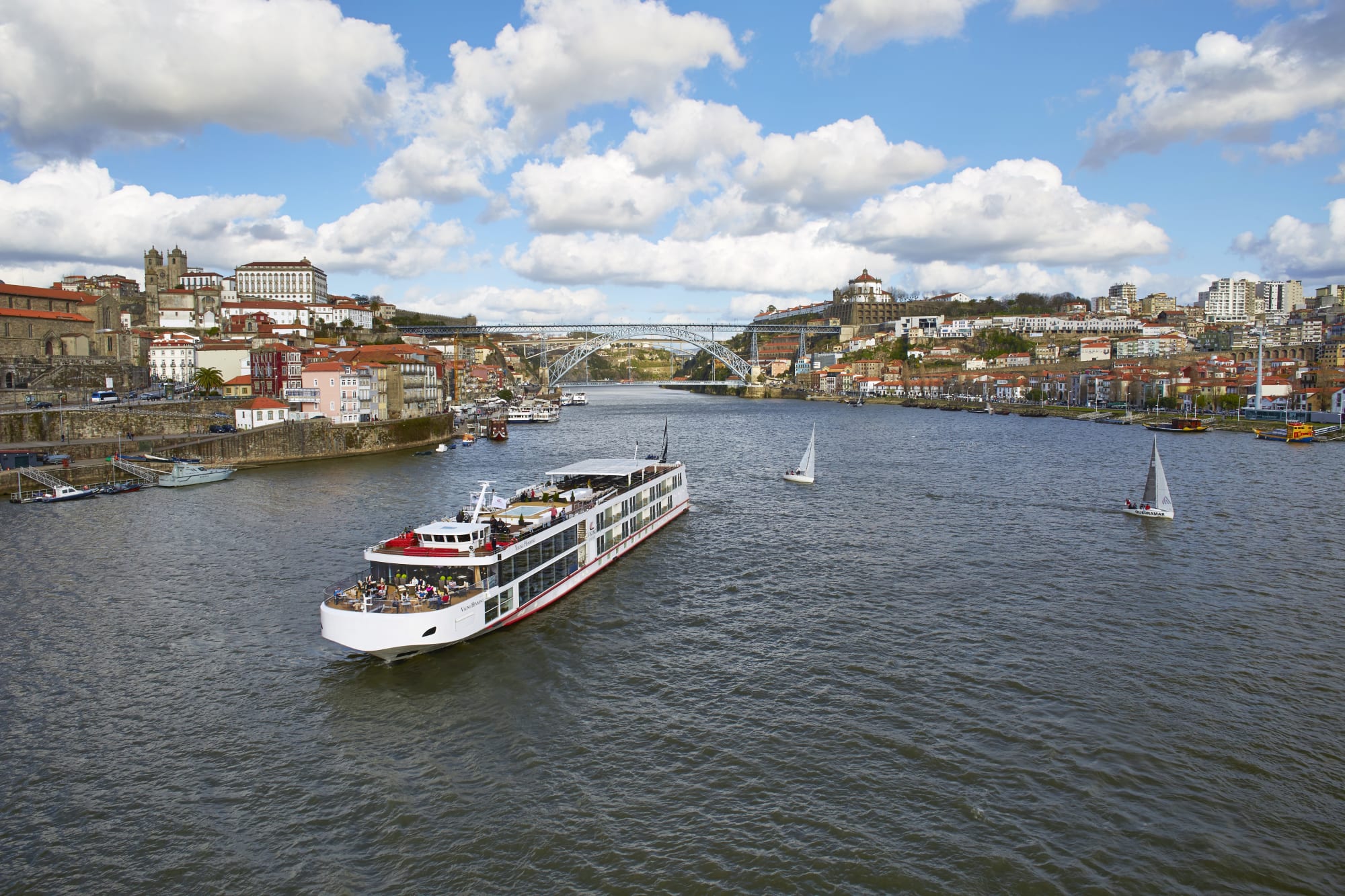 A modern river cruise ship navigates the Douro River in Porto, with the iconic Dom Luis I Bridge and historic city architecture in the background.