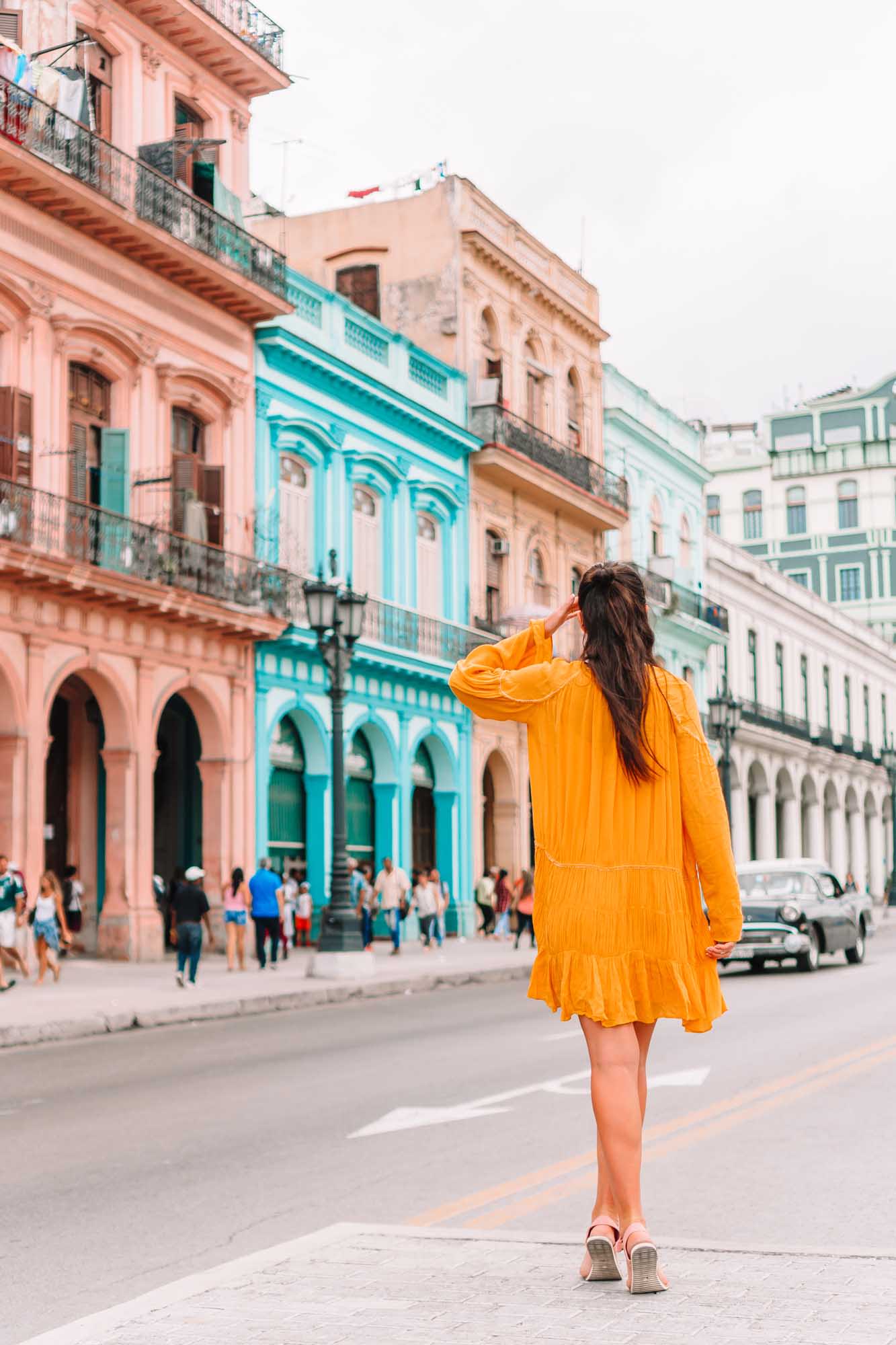 A young woman wearing a vibrant yellow dress stands on a colorful street in Old Havana, Cuba, gazing at the beautifully preserved colonial architecture featuring pastel pink, turquoise, and cream-colored buildings with ornate balconies and arched windows.