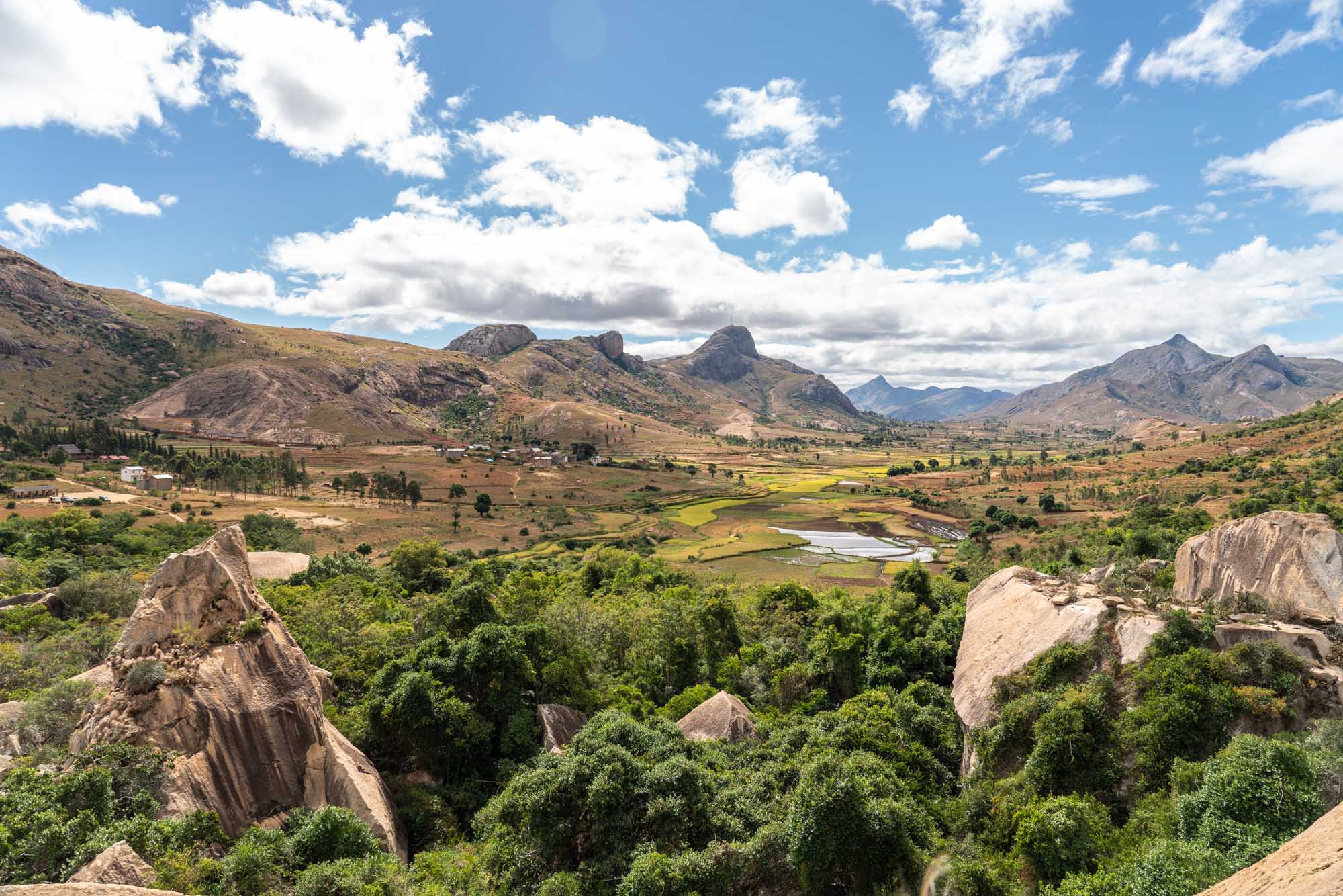 A panoramic view of the Anja Community Reserve in Madagascar, featuring distinctive large granite boulders scattered across a lush green landscape with rice paddies, distant mountains, and a winding river. The scene combines dramatic rocky outcrops with cultivated agricultural fields and natural vegetation under a partly cloudy sky.