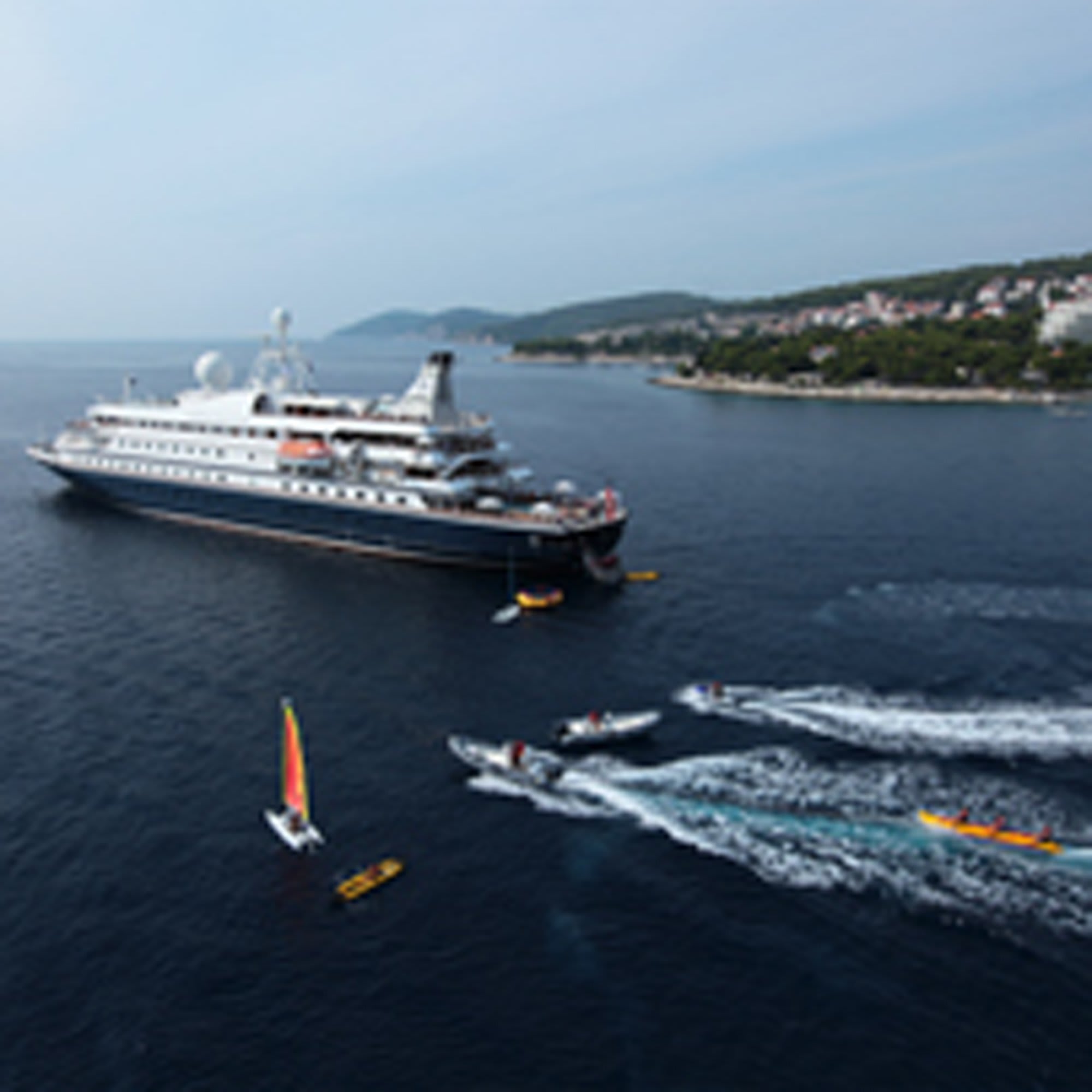 A large cruise ship anchored in calm Mediterranean waters with jet skis and water sports equipment