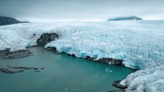 A stunning Arctic glacier descends steeply to meet turquoise glacial meltwater, with ice cliffs towering above rocky shores and mountains visible in the misty background. The striking contrast between brilliant blue ice, white snow, and the vibrant turquoise water showcases the raw beauty of a polar ice landscape, likely in Svalbard or similar Arctic destination.