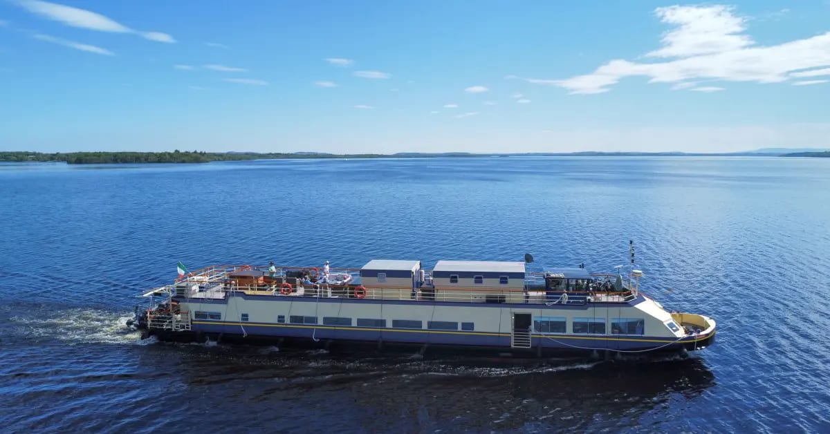 A multi-deck riverboat or lake cruise vessel anchored in calm blue waters with forested shoreline