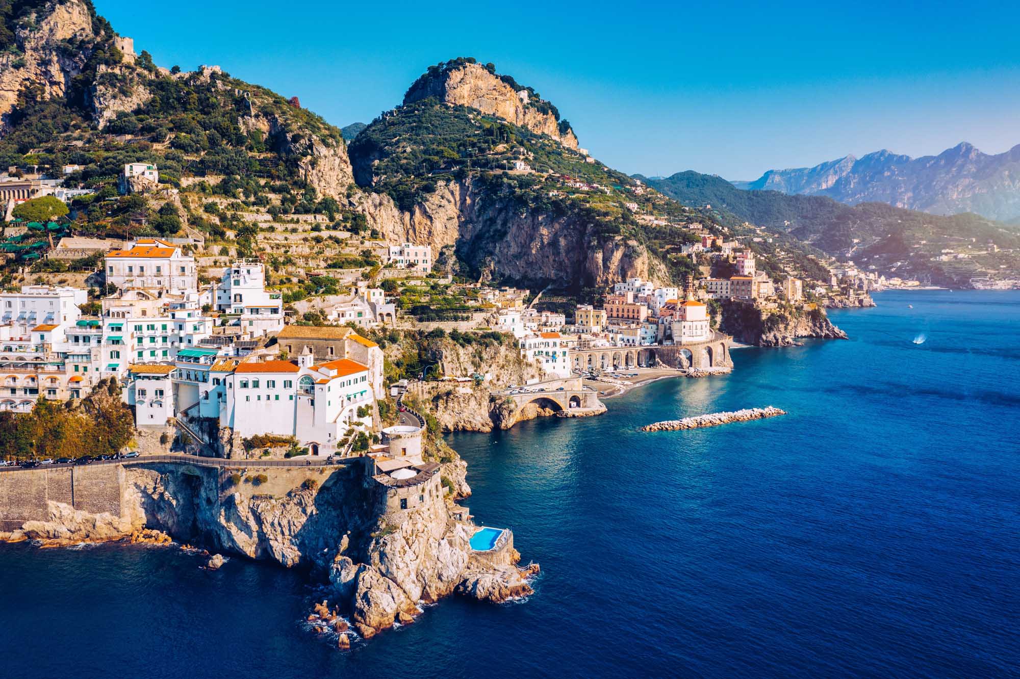 An aerial view of Atrani, a picturesque coastal village clinging to dramatic cliffsides along Italy's Amalfi Coast. Colorful pastel-hued buildings cascade down the rocky terrain toward crystal-clear turquoise Mediterranean waters, with the surrounding mountains and neighboring villages visible in the distance.