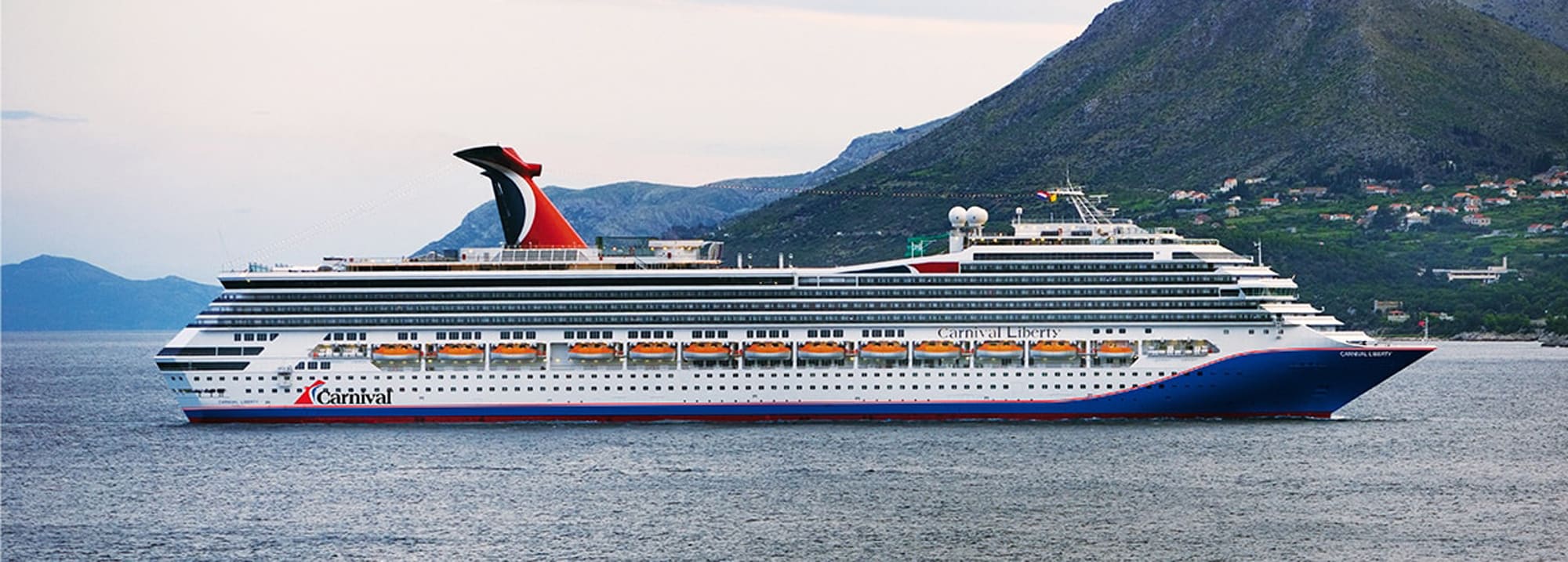 A large Carnival cruise ship sailing near a mountainous coastline with green hills and buildings