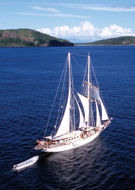 A stunning white-sailed tall ship glides through deep blue tropical waters with lush, forested
