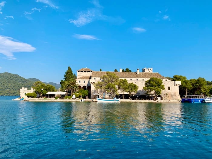 A stunning coastal fortress situated on a rocky island surrounded by crystal-clear turquoise Mediterranean waters, featuring classical stone architecture with towers and fortified walls, framed by lush green hillsides and traditional fishing boats nearby.