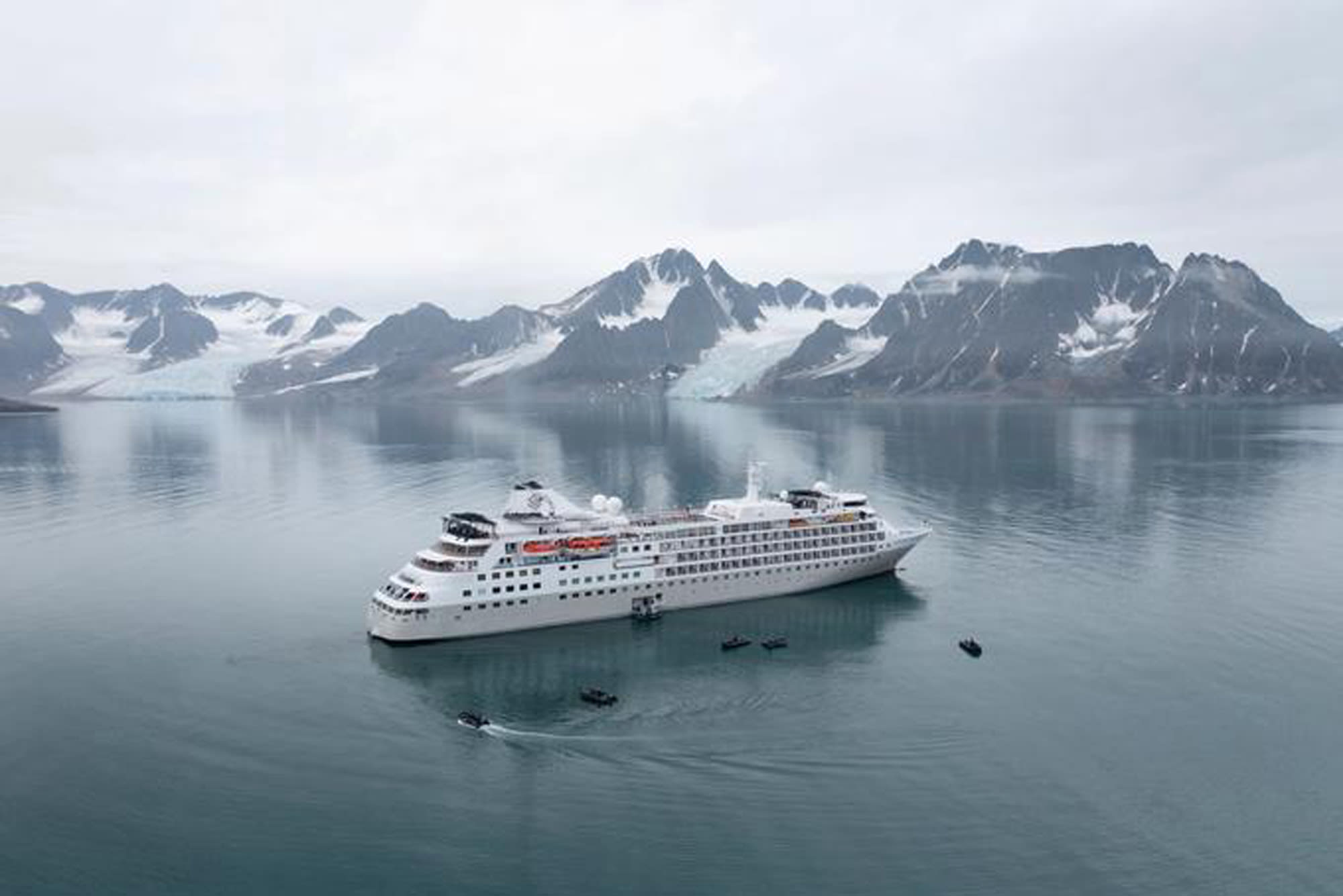 A large white expedition cruise ship anchors in calm, mirror-like Arctic waters with small tender