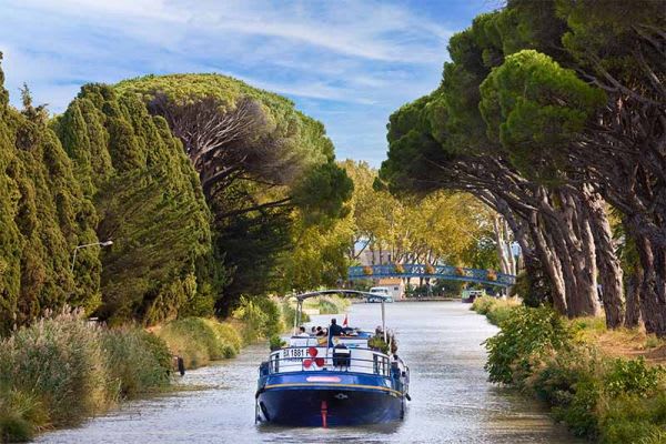 A blue and red commercial barge navigates through a scenic European canal lined with tall