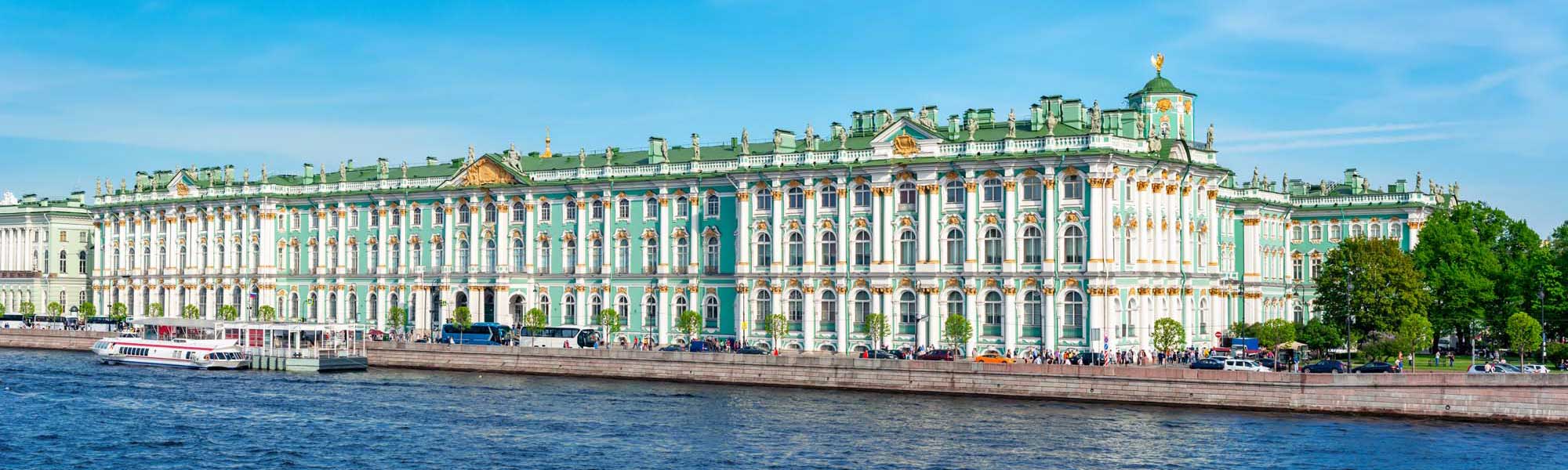 The magnificent Winter Palace in St. Petersburg, Russia, displaying its iconic turquoise and white Baroque façade with golden ornamental details, as viewed from the Neva River. The grand 18th-century imperial residence features multiple stories of arched windows, decorative moldings, and a distinctive green roof punctuated by ornamental spires.