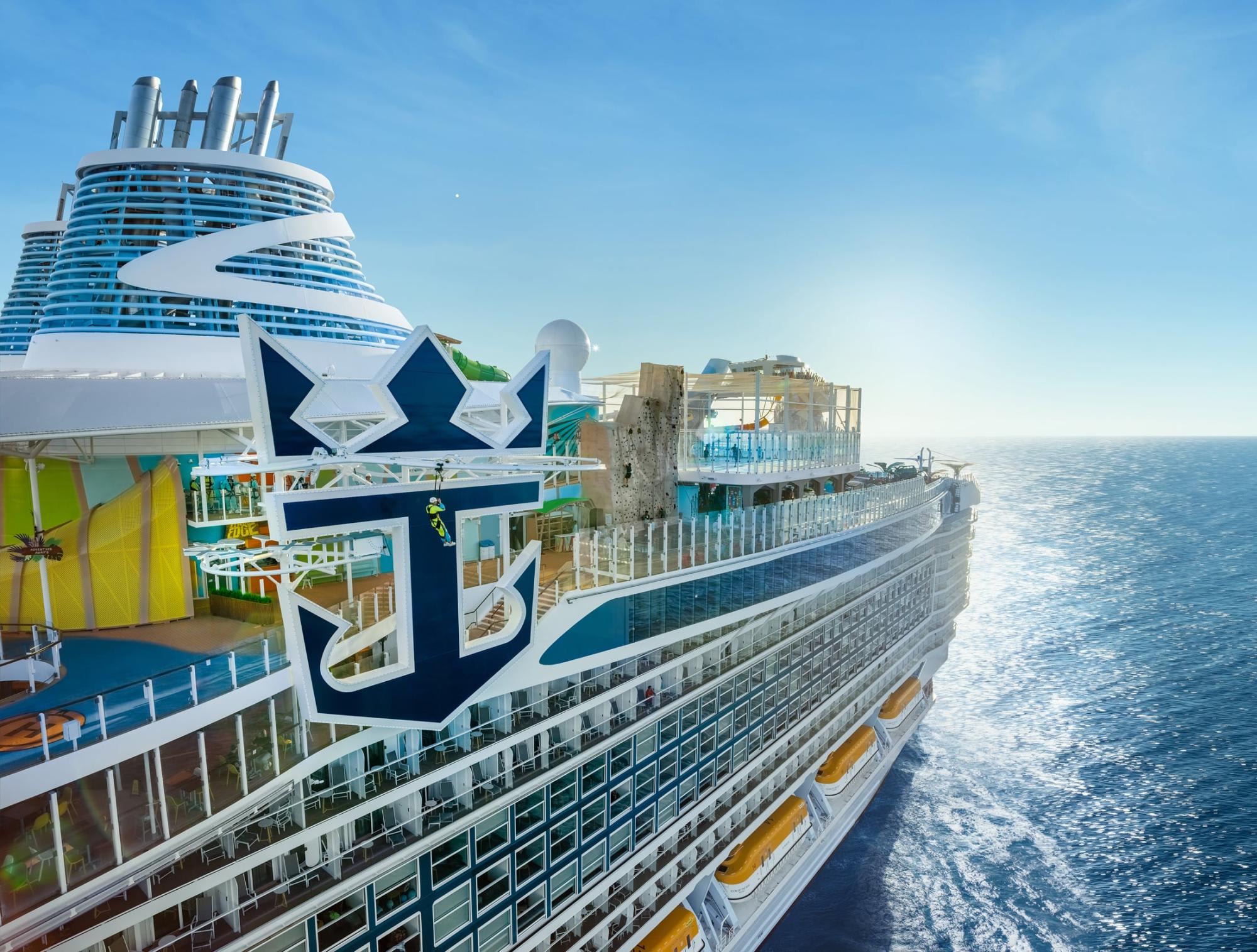 A modern cruise ship with distinctive blue and white livery featuring a large anchor emblem on its hull, photographed from the side while at sea. The ship displays multiple decks with balconies, colorful amenities including yellow waterslides, and multiple smokestack funnels, representing contemporary cruise industry design and luxury ocean travel.