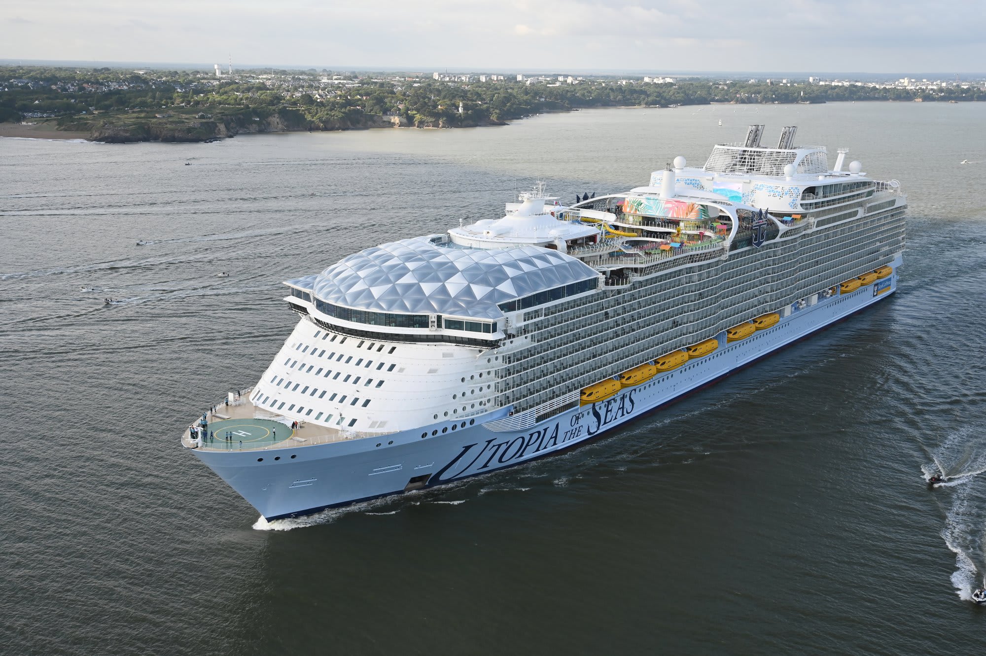 A large modern cruise ship with distinctive light blue hull and white superstructure anchored in coastal waters, featuring a prominent geodesic dome structure on the forward deck and colorful entertainment areas visible amidships. The ship is positioned near a residential coastline with tree-lined shores and distant urban development visible in the background.