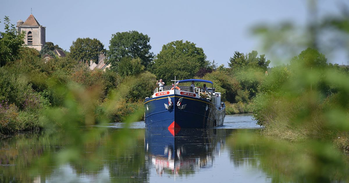 A blue and black river cruise barge navigates a peaceful inland waterway lined with lush green vegetation, with a charming church featuring a distinctive bell tower visible in the background among trees. This scene captures the serene experience of river cruising through the European countryside.