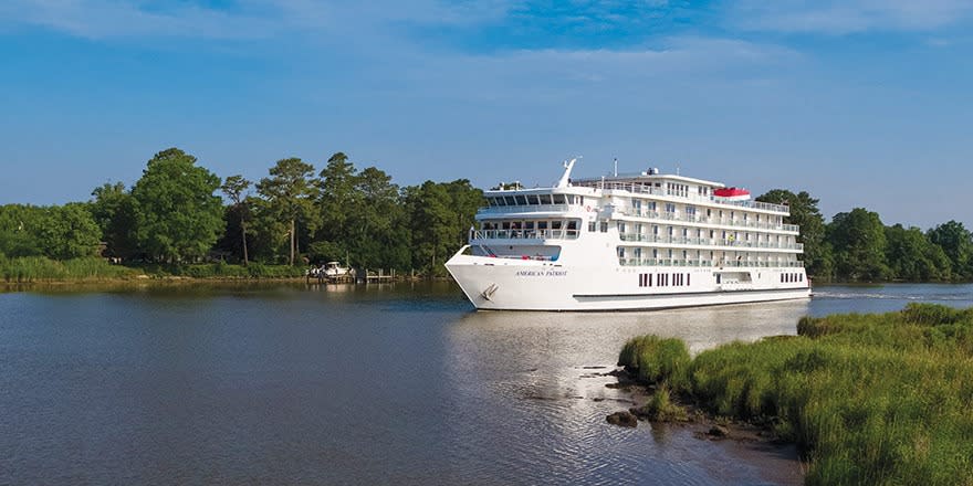 A large multi-deck river cruise ship operated by American Cruise Lines navigates a scenic waterway