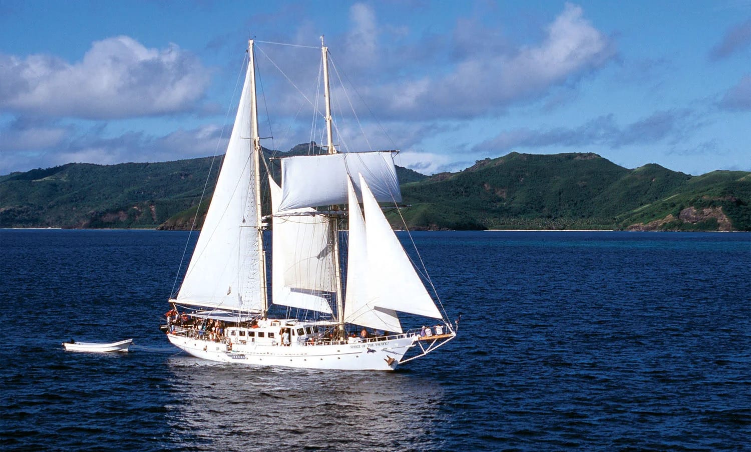 A stunning white two-masted sailing ship anchors in crystal-clear tropical waters, with lush