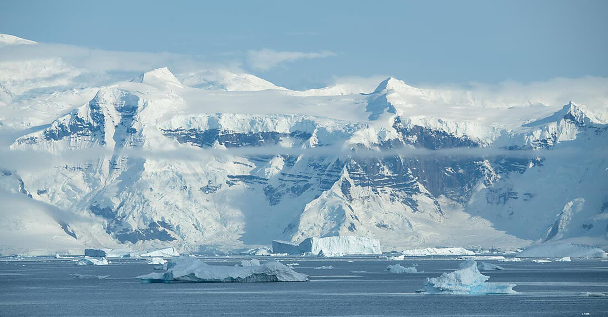A stunning polar landscape featuring towering snow-covered mountains rising dramatically from the sea, with numerous icebergs floating in the foreground. The pristine white peaks are partially shrouded in mist, creating a serene and majestic Antarctic seascape under clear blue skies.