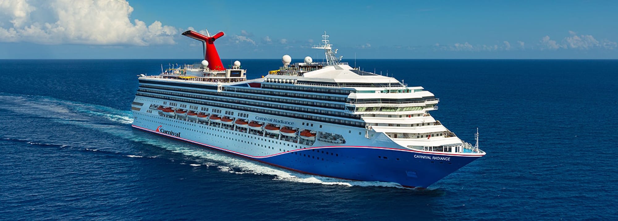 A large white and blue Carnival cruise ship sailing on open blue ocean waters with fluffy white