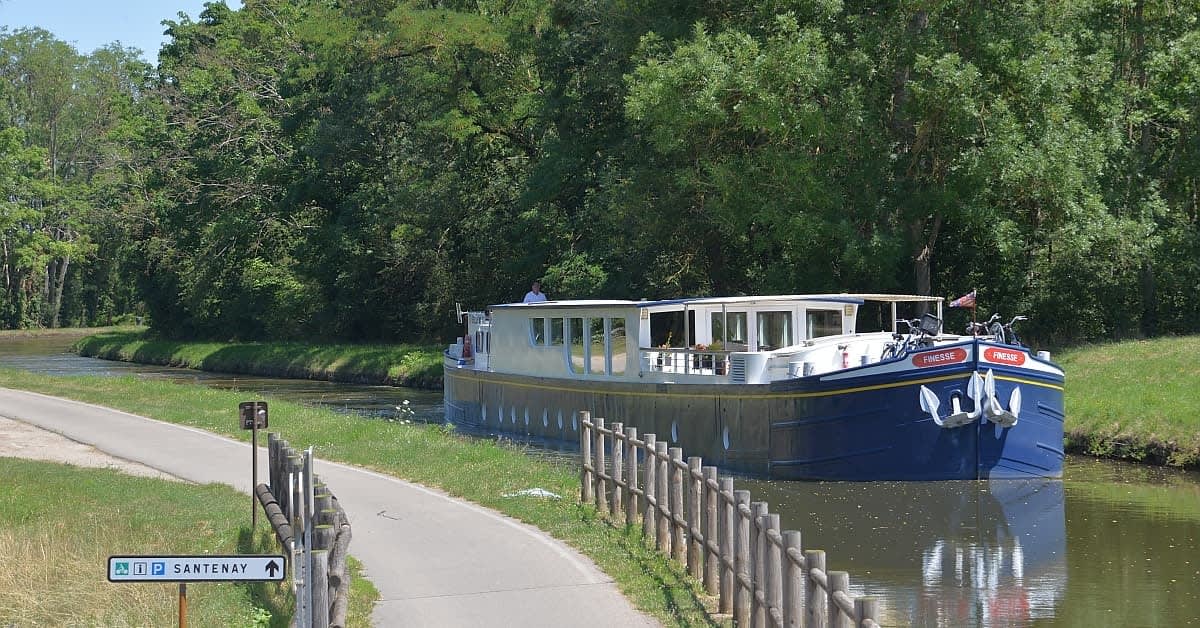 A modern river cruise vessel moored along a scenic European canal lined with lush green vegetation